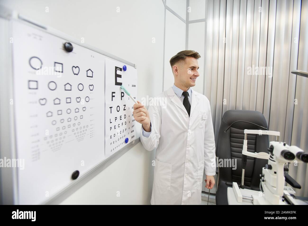 Portrait of smiling young ophthalmologist pointing at vision chart while checking eyesight of unrecognizable patient, copy space Stock Photo