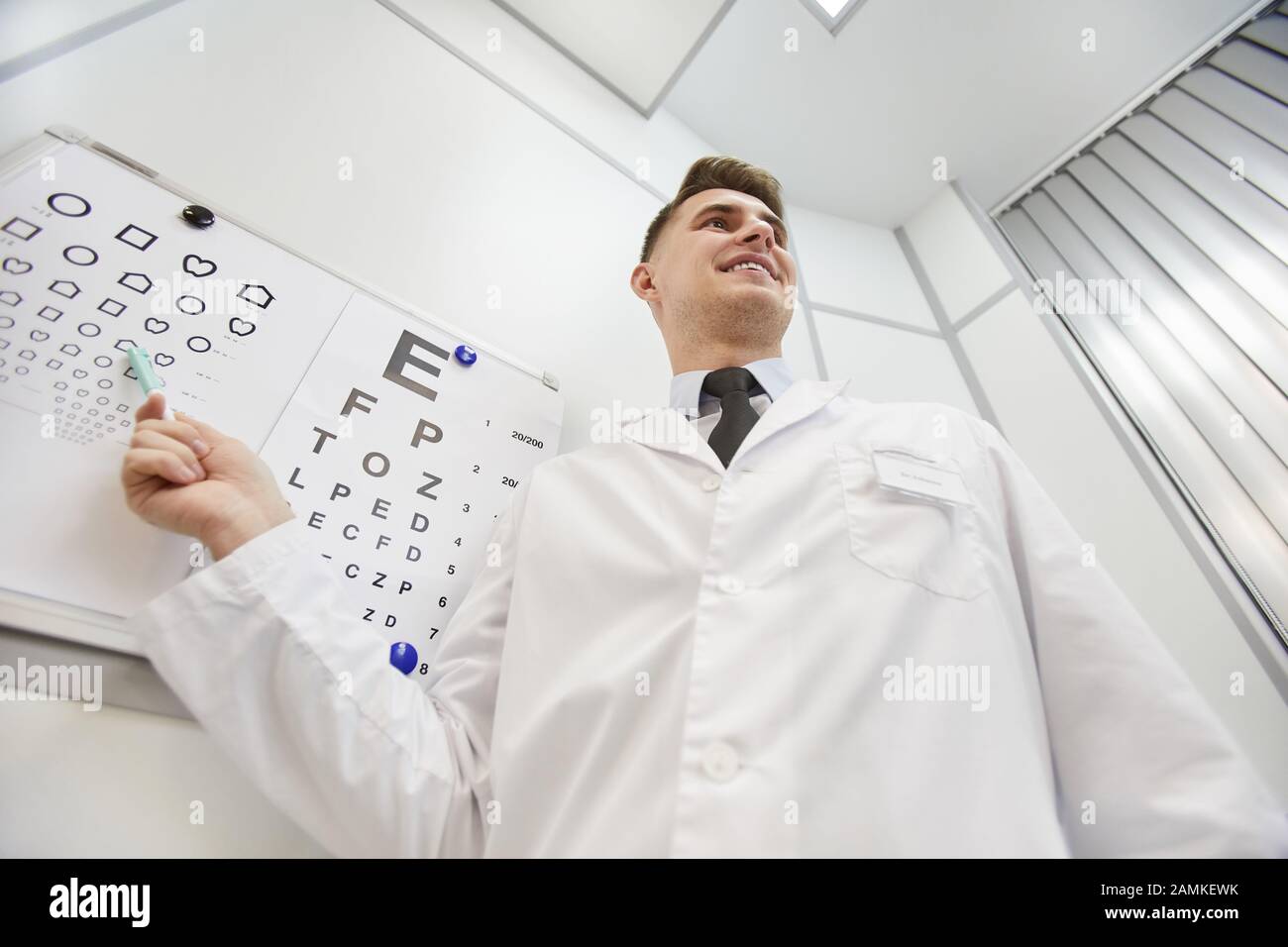 Low angle portrait of smiling young ophthalmologist pointing at vision chart while checking eyesight of unrecognizable patient, copy space Stock Photo