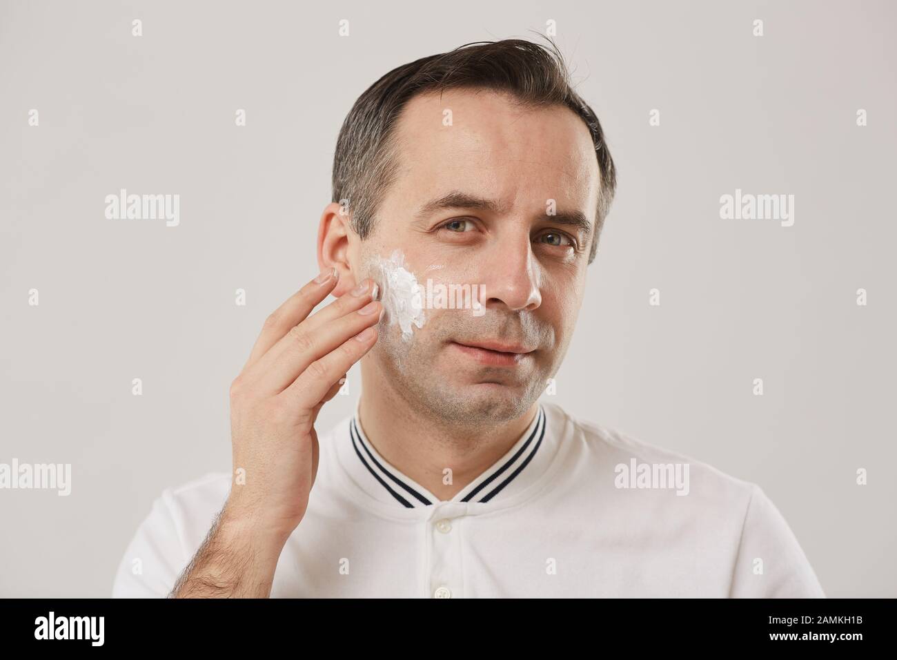 Head and shoulders portrait of middle aged man putting on shaving cream during morning routine standing against white background Stock Photo