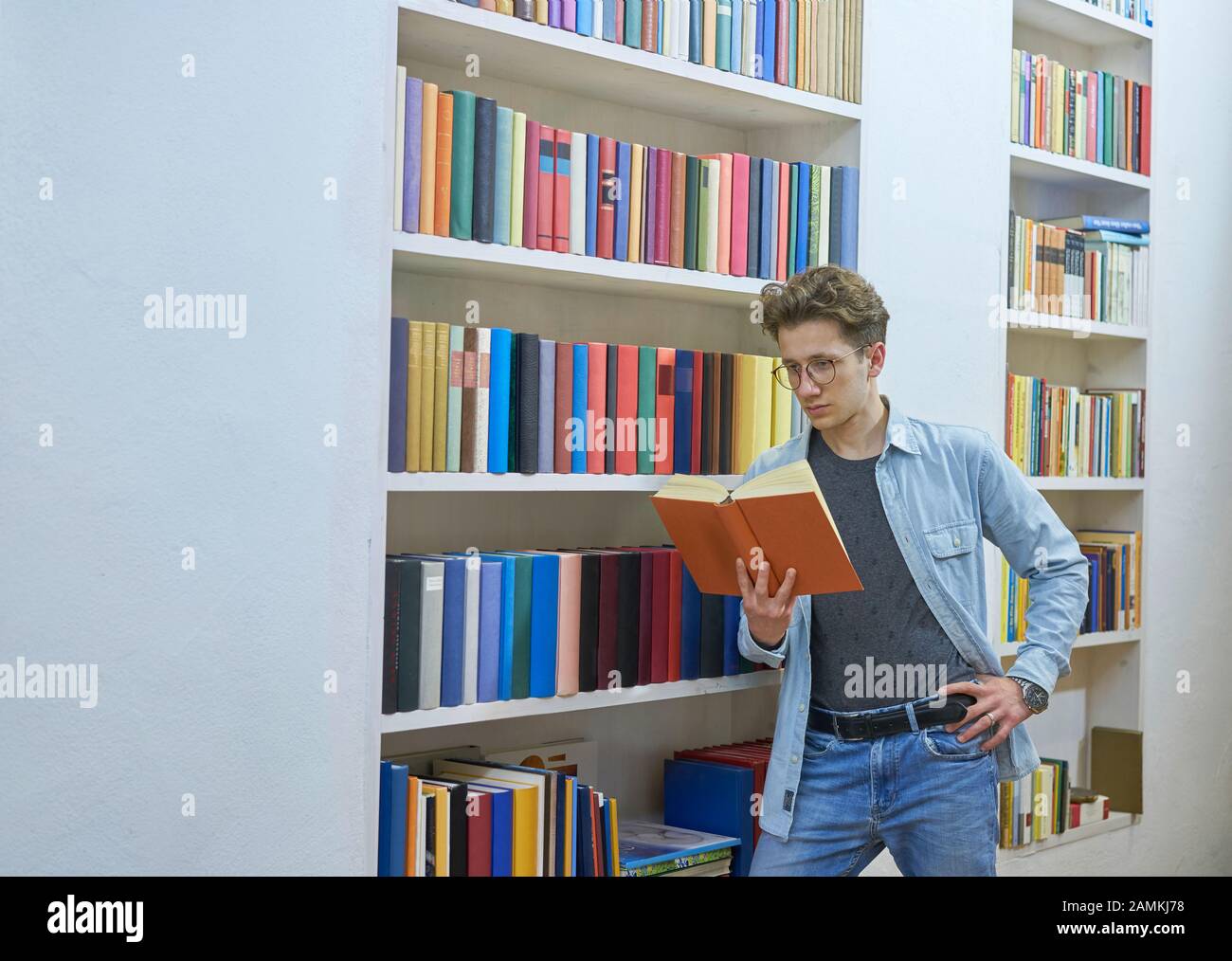 Young man reading a book while leaning on a bookshelf with colorful books in a library. Stock Photo