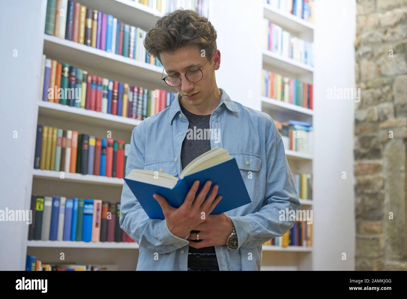 Young student with curls focussed on reading a book and standing  next to a bookshelf Stock Photo