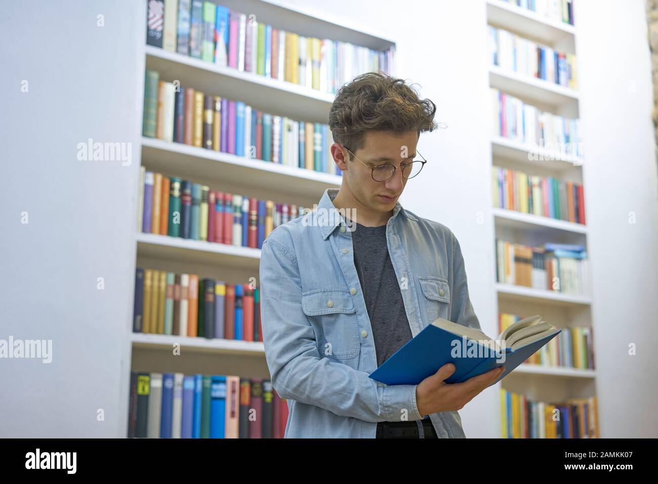 Young handsome man reading a book in a library with big bookshelves in the background. Stock Photo