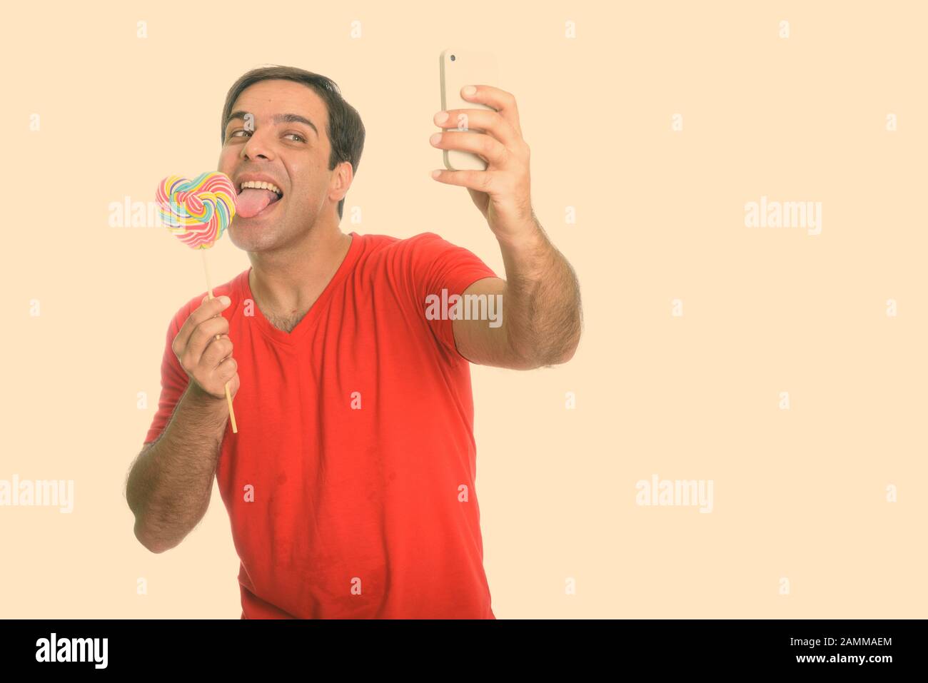 Happy handsome Persian man taking selfie while licking heart shaped lollipop Stock Photo