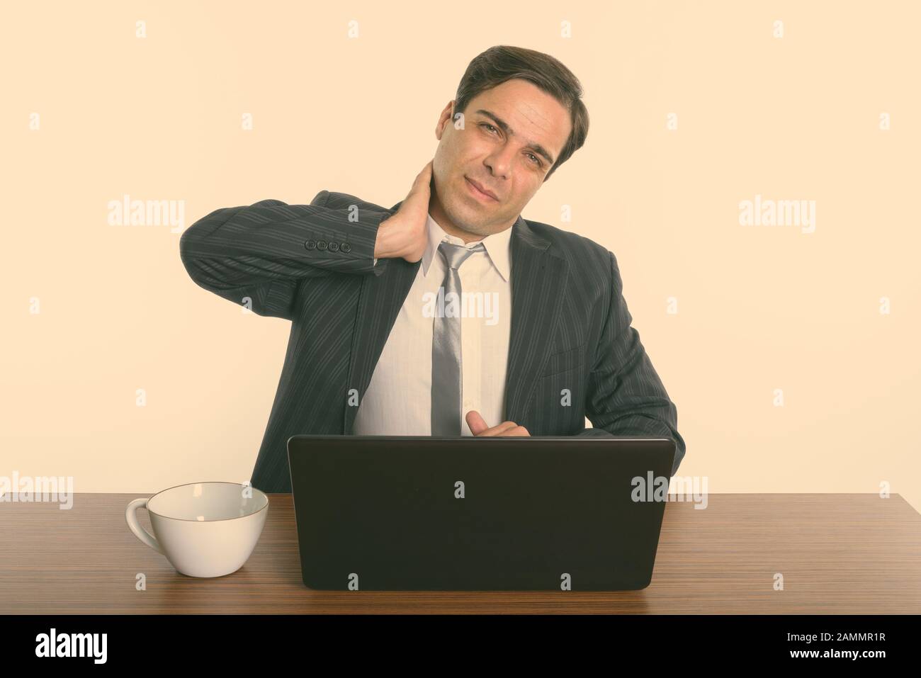 Stressed Persian businessman sitting while having neck pain with laptop on wooden table Stock Photo