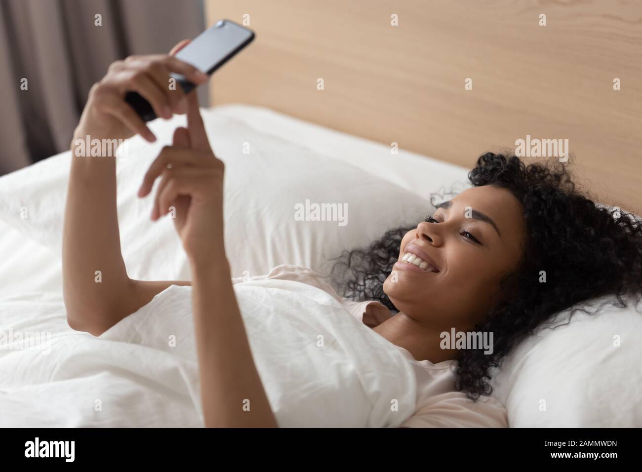 African woman lying down in bed with smartphone in morning Stock Photo