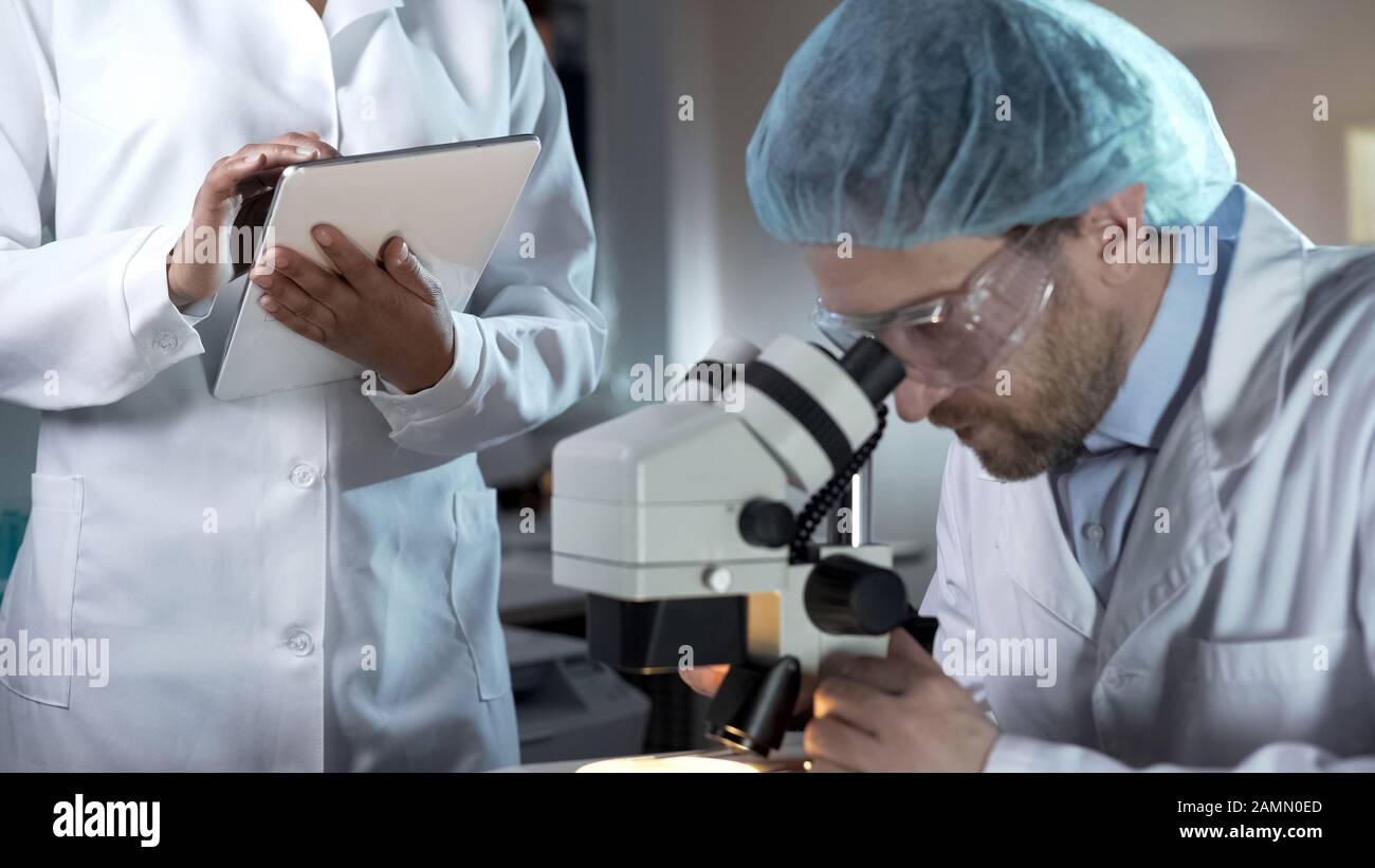 Researcher working with microscope, assistant recording comments on tablet Stock Photo