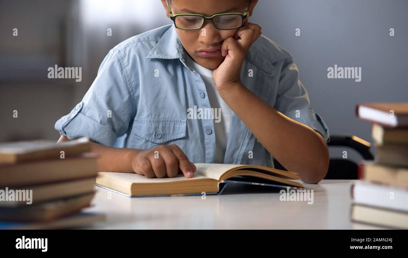 Smart boy in glasses sitting in library reading books, educational literature Stock Photo