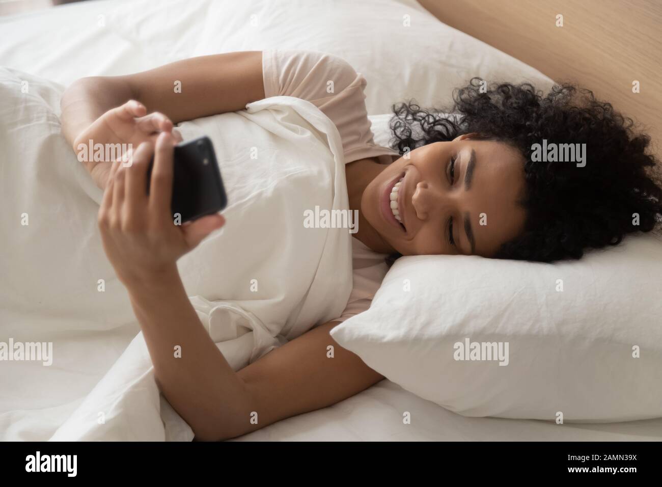 African woman lying in bed holding smartphone looking at screen Stock Photo