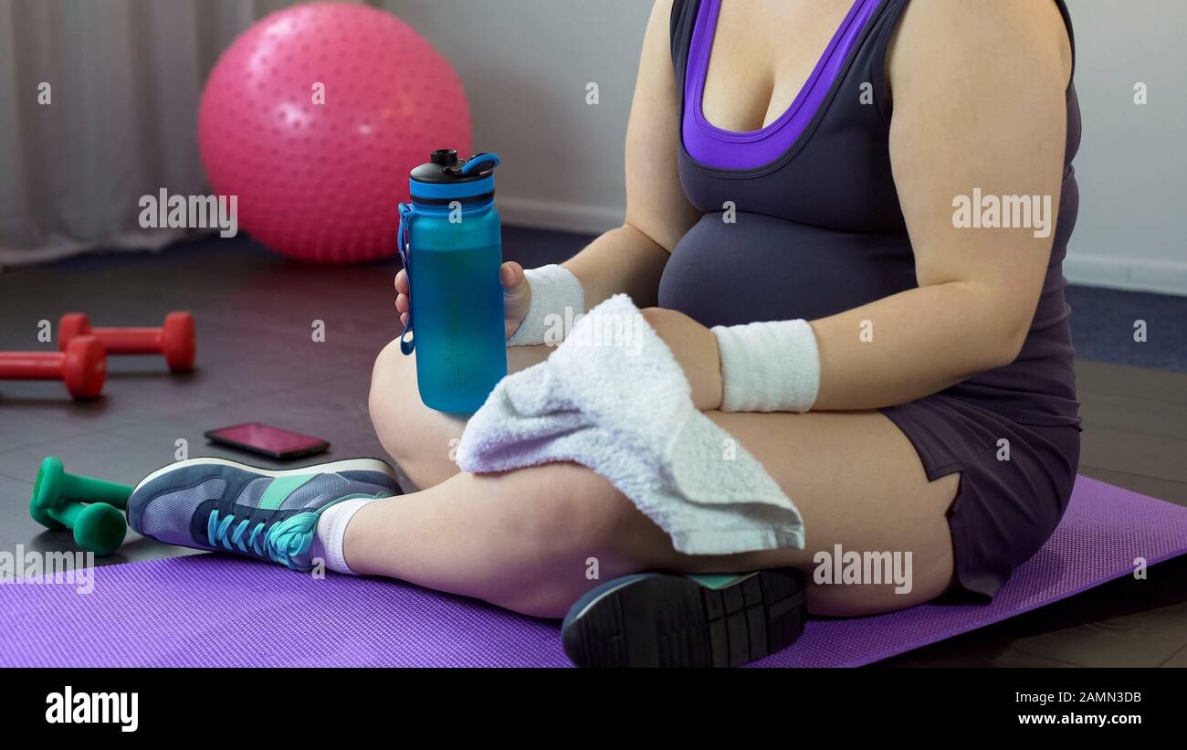 Plump lady drinking water and restoring breath after exhausting home workout Stock Photo