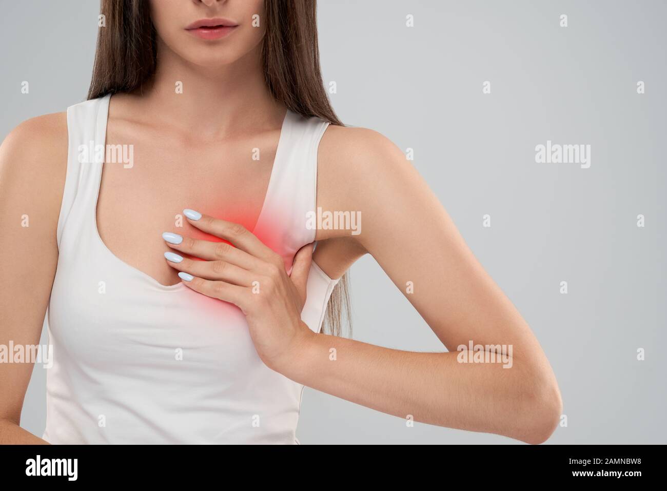 Clolse up of incognito caucasian woman in white shirt posing over gray isolated background and touching chest with hand because of heartache. Front crop of brunette suffering from pain in heart. Stock Photo