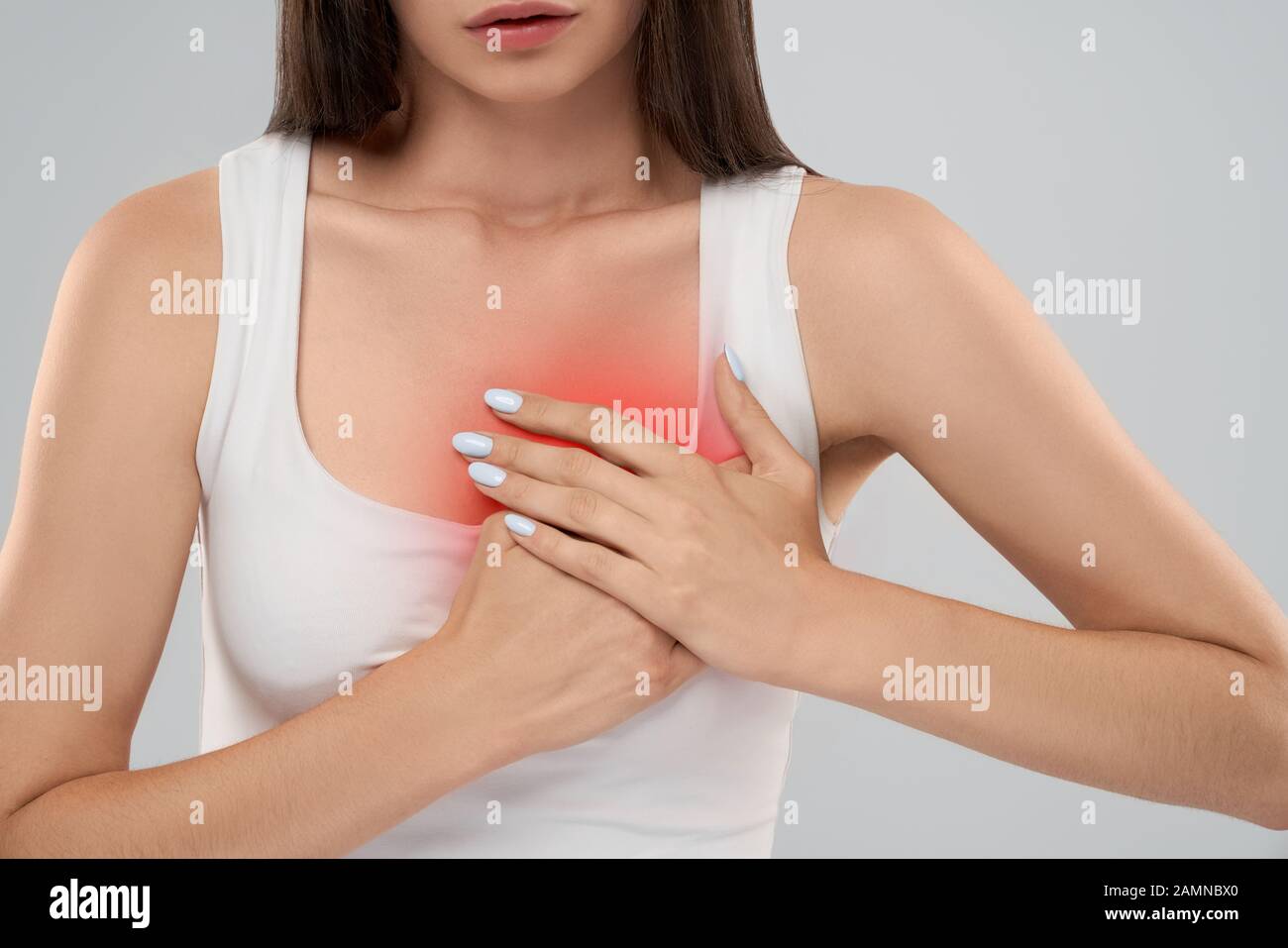 Clolse up of incognito caucasian woman in white shirt posing over gray isolated background and touching chest with two hands because of heartache. Front crop of brunette suffering from pain in heart. Stock Photo