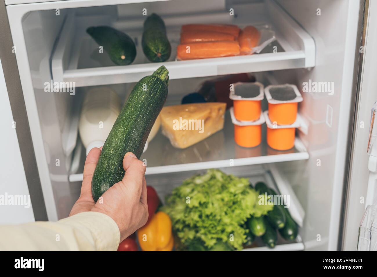 cropped view of man holding cucumber near open fridge full of food Stock Photo