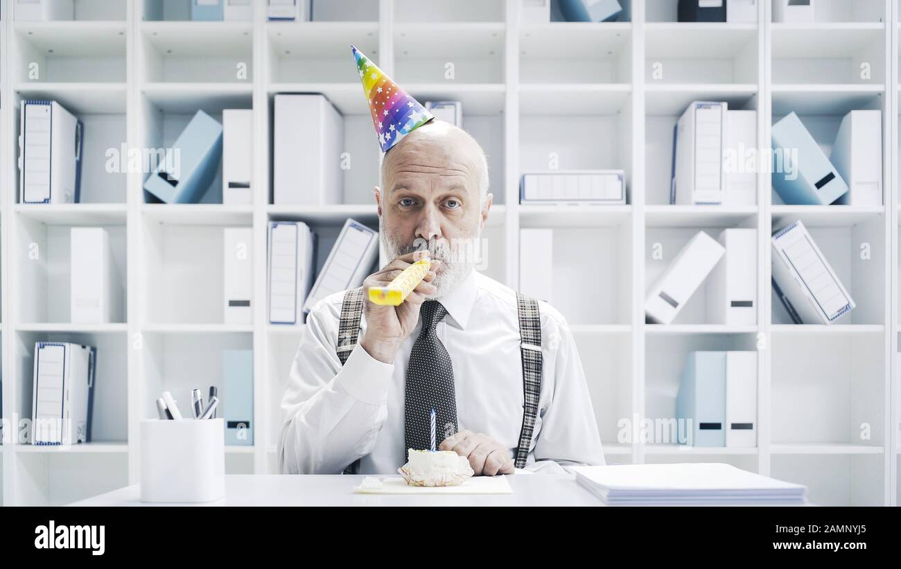 Senior businessman having a sad lonely birthday in the office, he is wearing a party hat and having a small cake alone Stock Photo