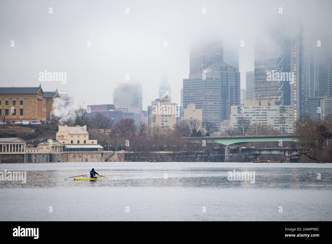 A single rower in a scull rows on a river in front of a mist-covered cityscape. Stock Photo