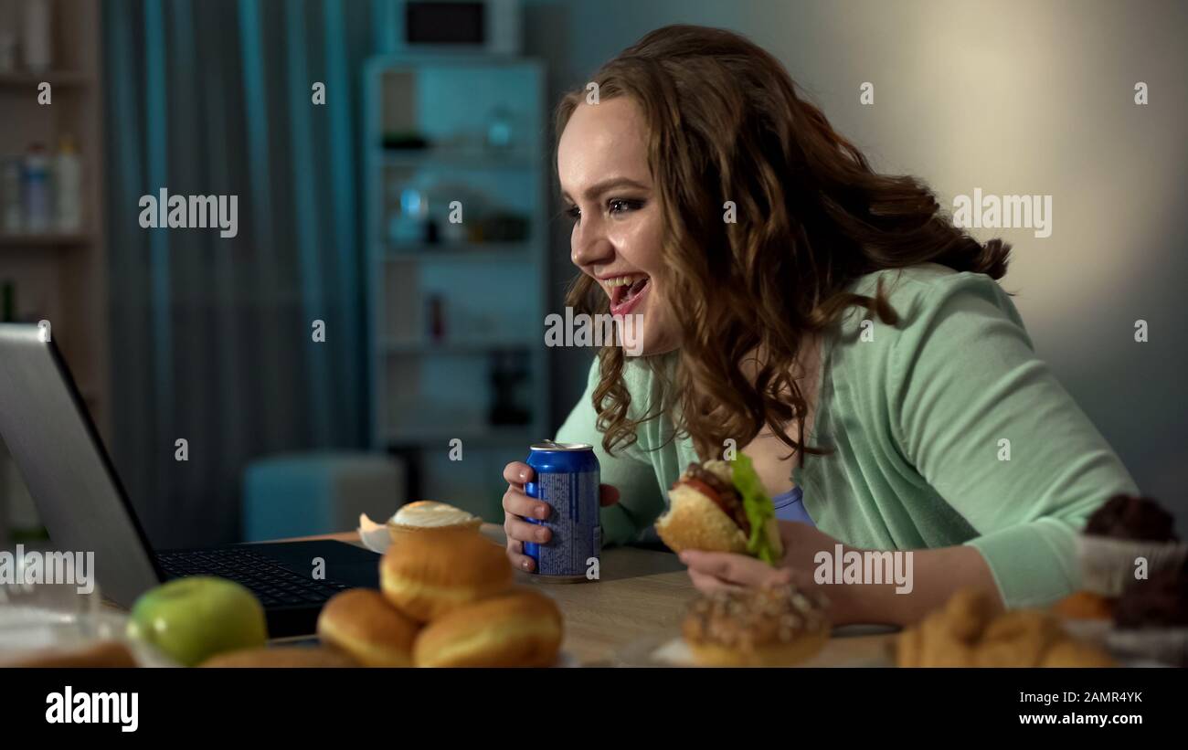 Bad-mannered obese girl laughing at show on laptop, eating unhealthy food Stock Photo