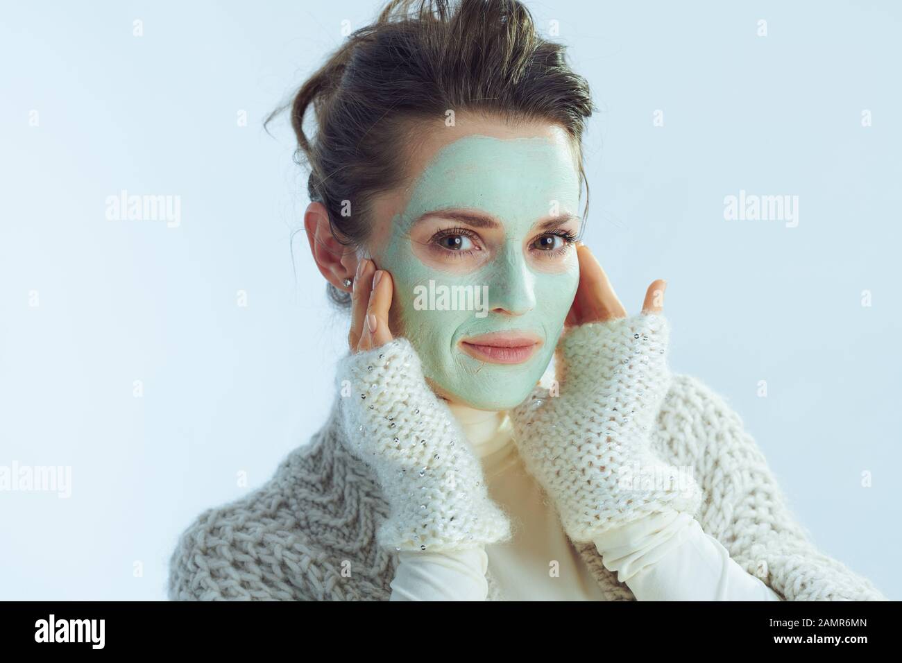 Portrait of modern 40 years old woman in roll neck sweater and cardigan with green facial mask touching face isolated on winter light blue background. Stock Photo