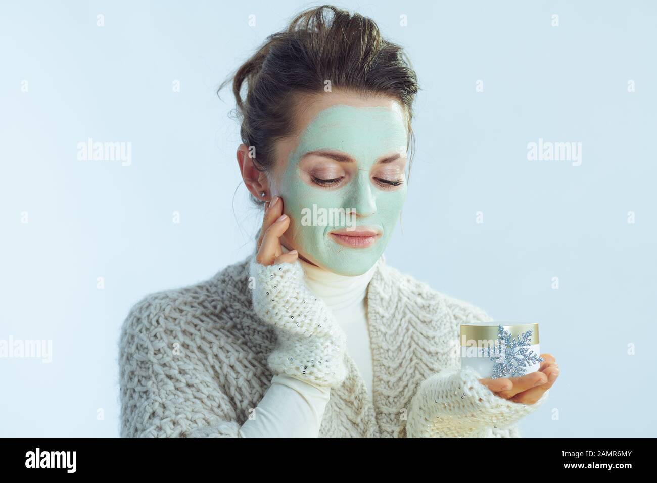 stylish woman in roll neck sweater and cardigan with green facial mask touching face and holding cosmetic jar and snowflake on winter light blue backg Stock Photo