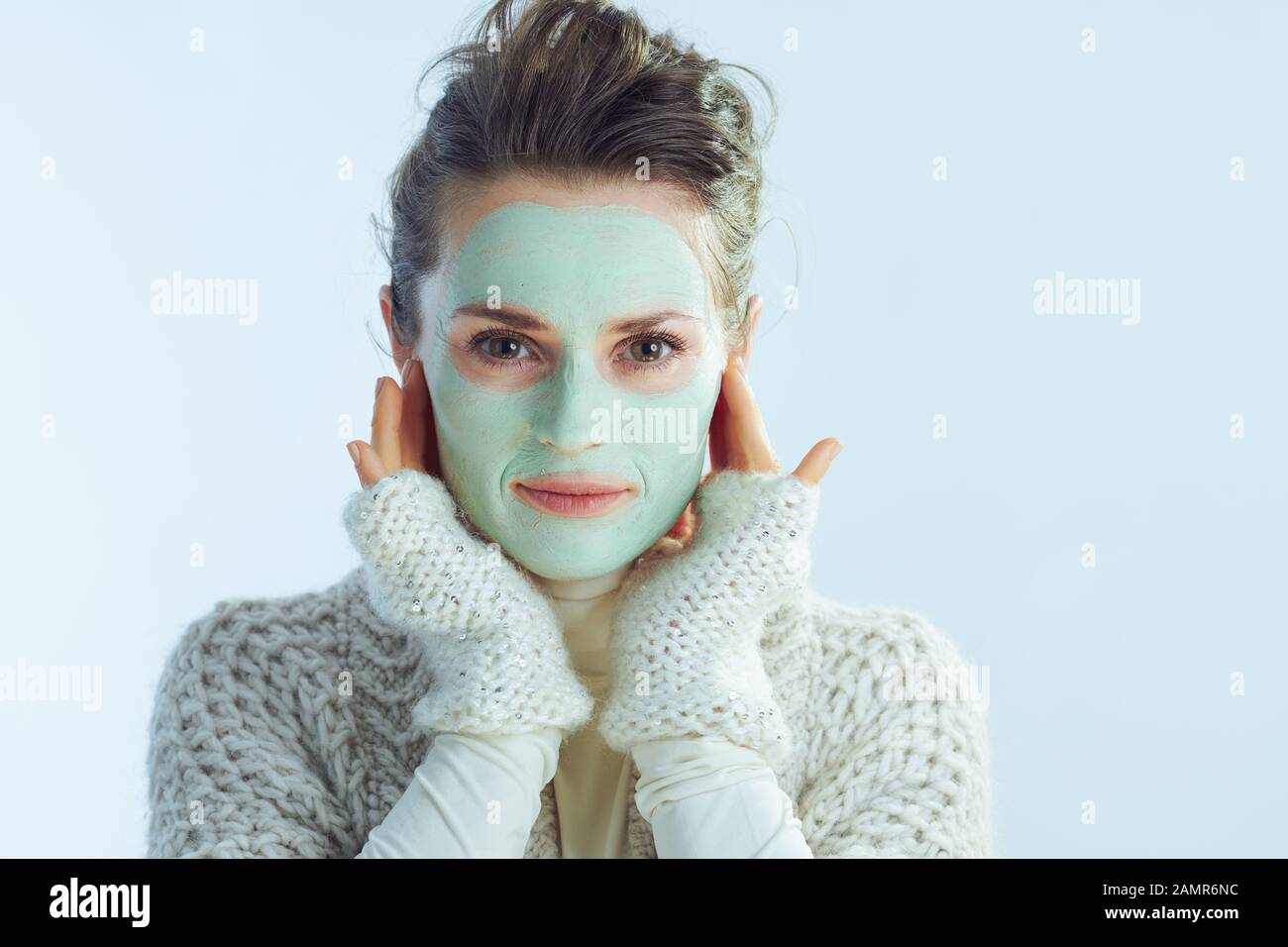 Portrait of stylish 40 years old housewife in roll neck sweater and cardigan with green facial mask touching face on winter light blue background. Stock Photo