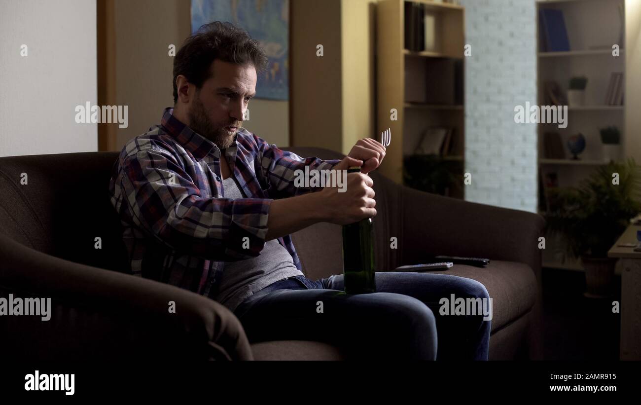 Depressed male sitting on sofa in dark room, opening beer bottle with fork Stock Photo