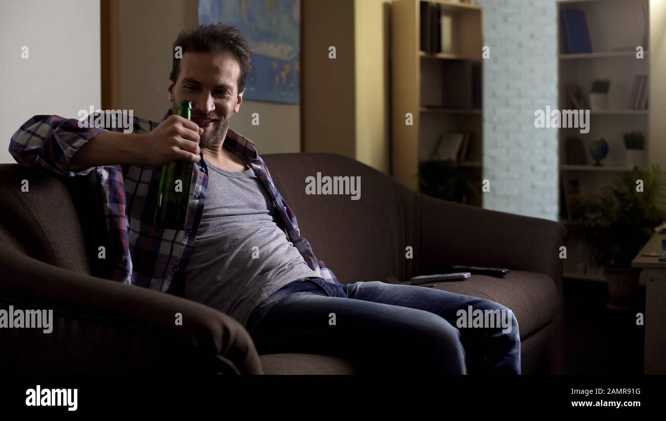 Drunk alcoholic enjoying tasty bottled beer, sitting on couch at home, bad habit Stock Photo
