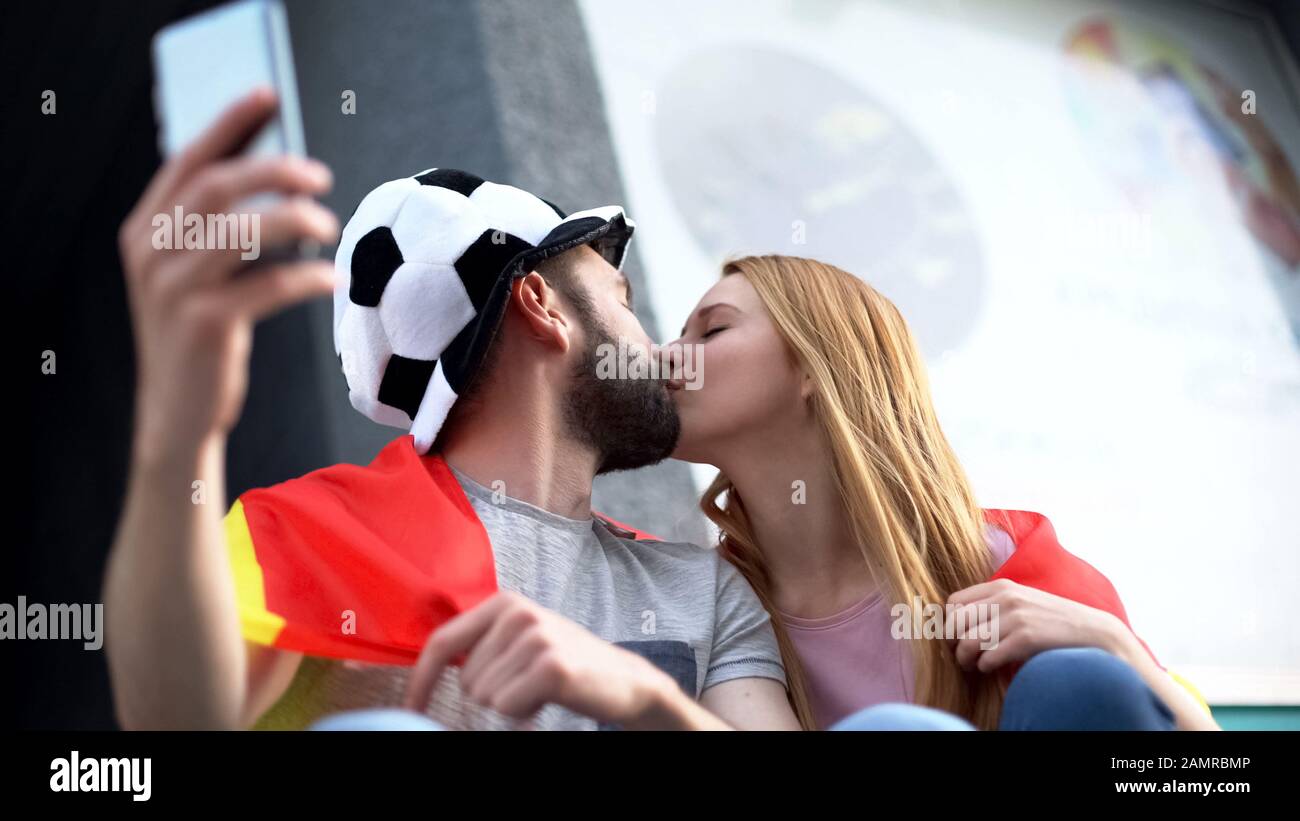 Couple of football fans kissing, taking selfie on smartphone, young