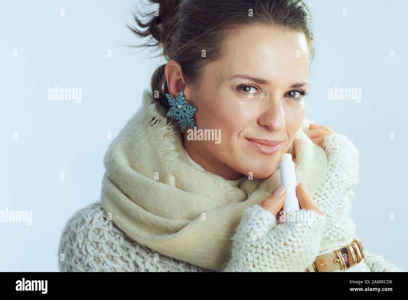 Portrait of elegant housewife in roll neck sweater and cardigan using lip balm as winter lip care on winter light blue background. Stock Photo