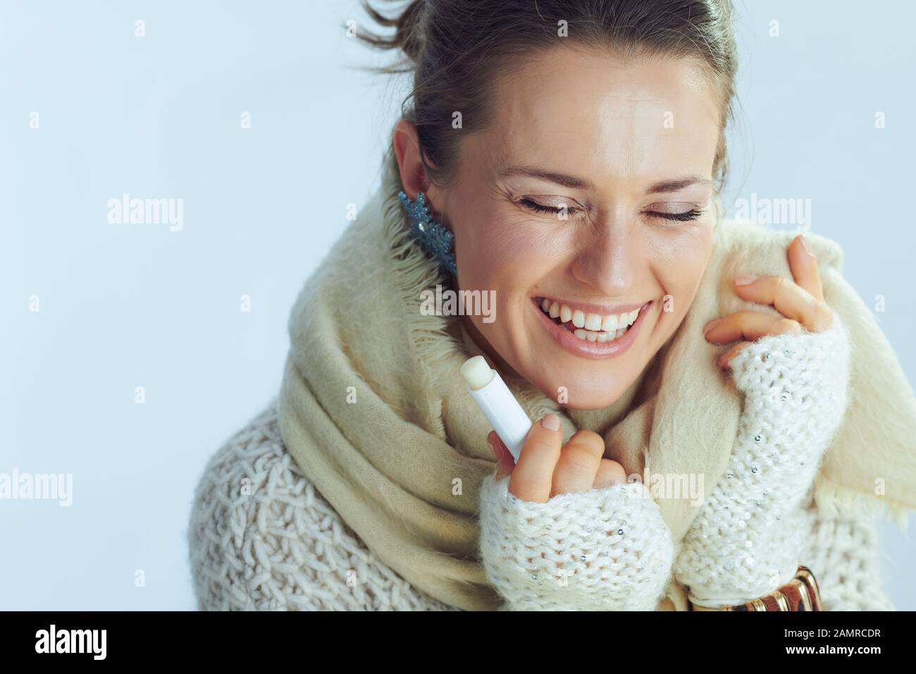happy stylish middle age woman in roll neck sweater and cardigan with lip balm as winter lip care on winter light blue background. Stock Photo