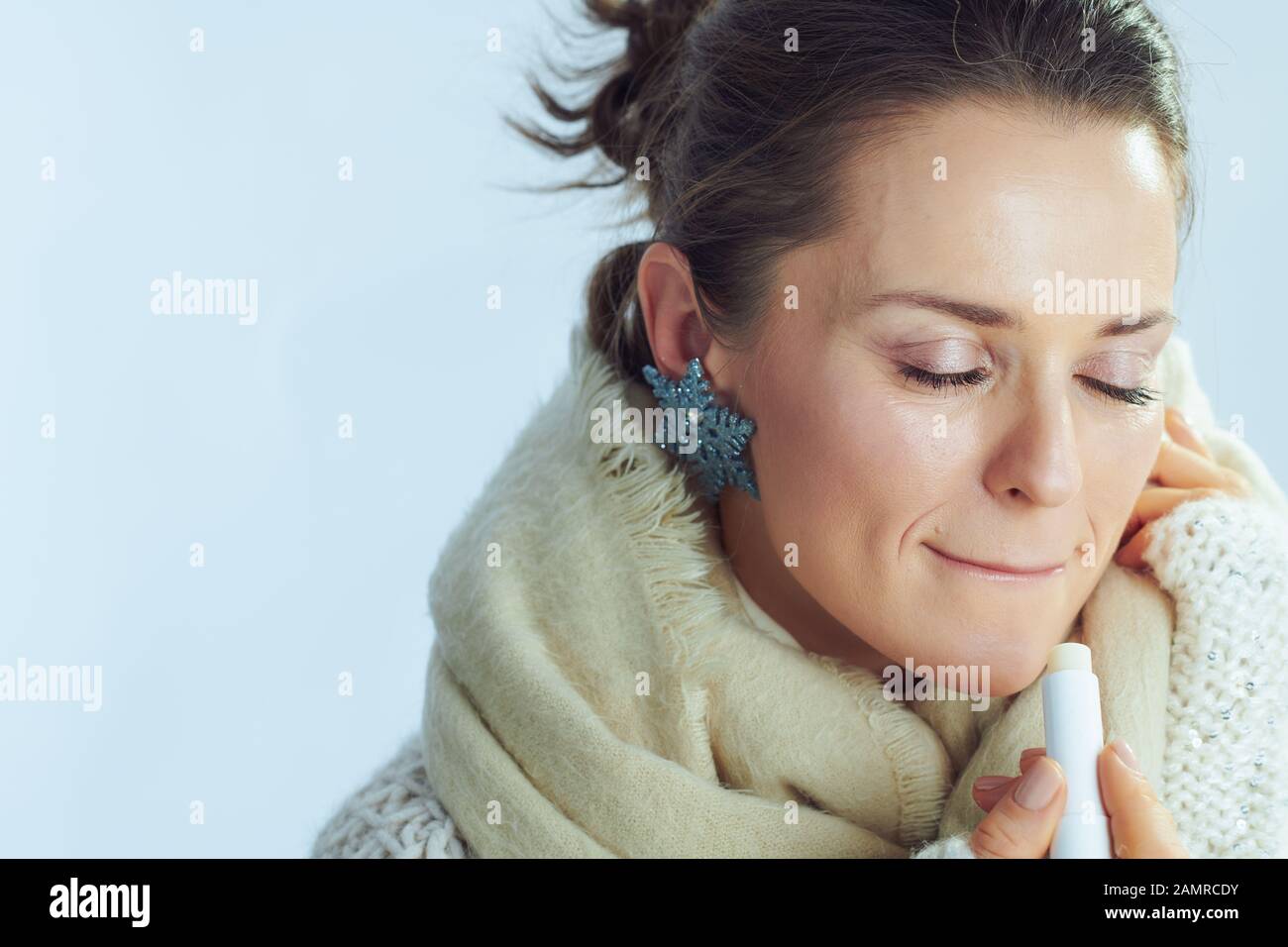 stylish 40 years old housewife in roll neck sweater and cardigan using lip balm as winter lip care against winter light blue background. Stock Photo