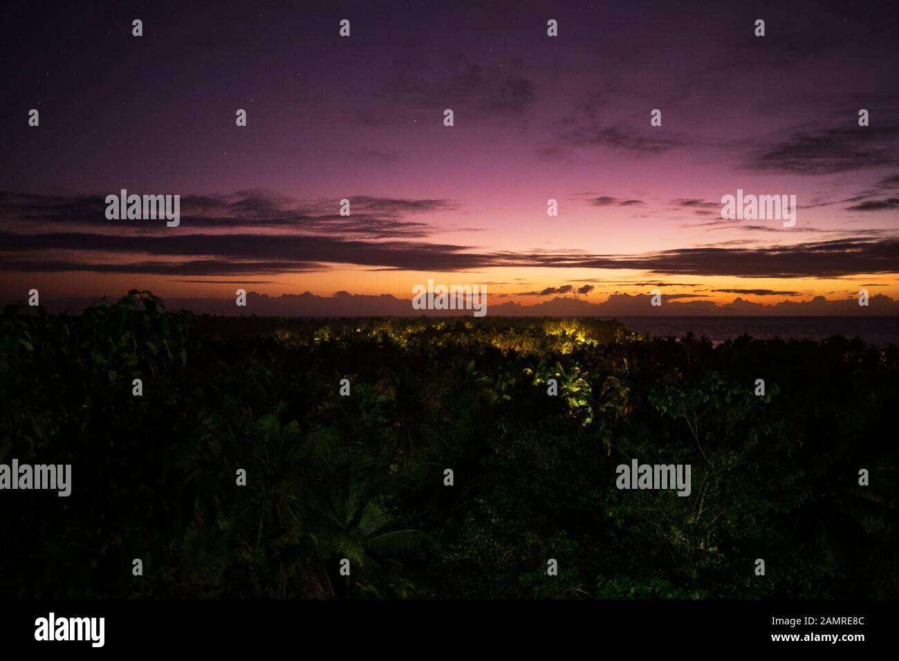 Stars from night sky fade into predawn color over tropical jungle lit by small village Stock Photo