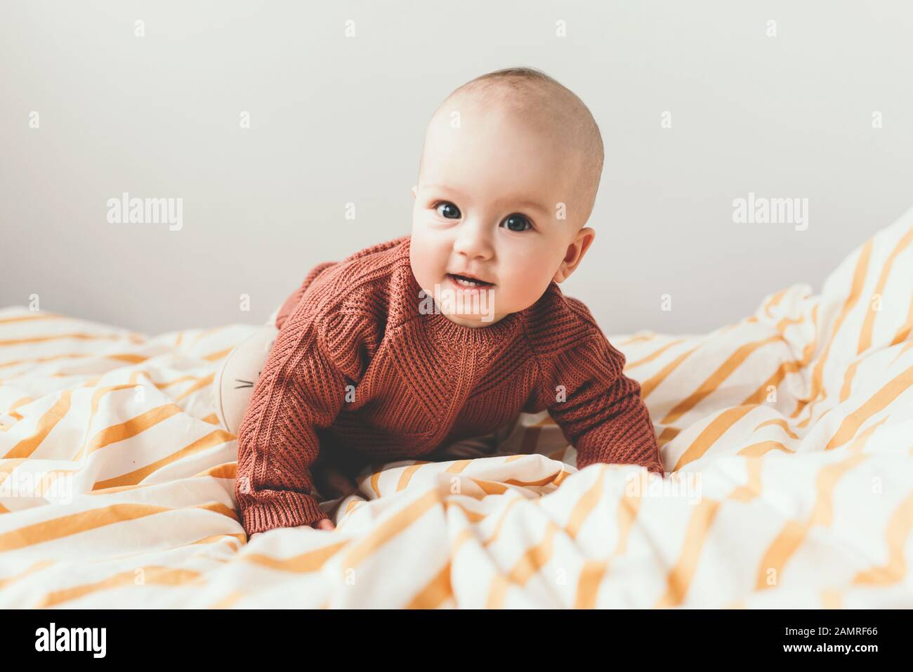 Beautiful little baby girl on the bed in a cozy brown sweater smiles. Concept of motherhood and childhood. Adorable six month old baby girl lying on the bad and looking into the camera. Stock Photo