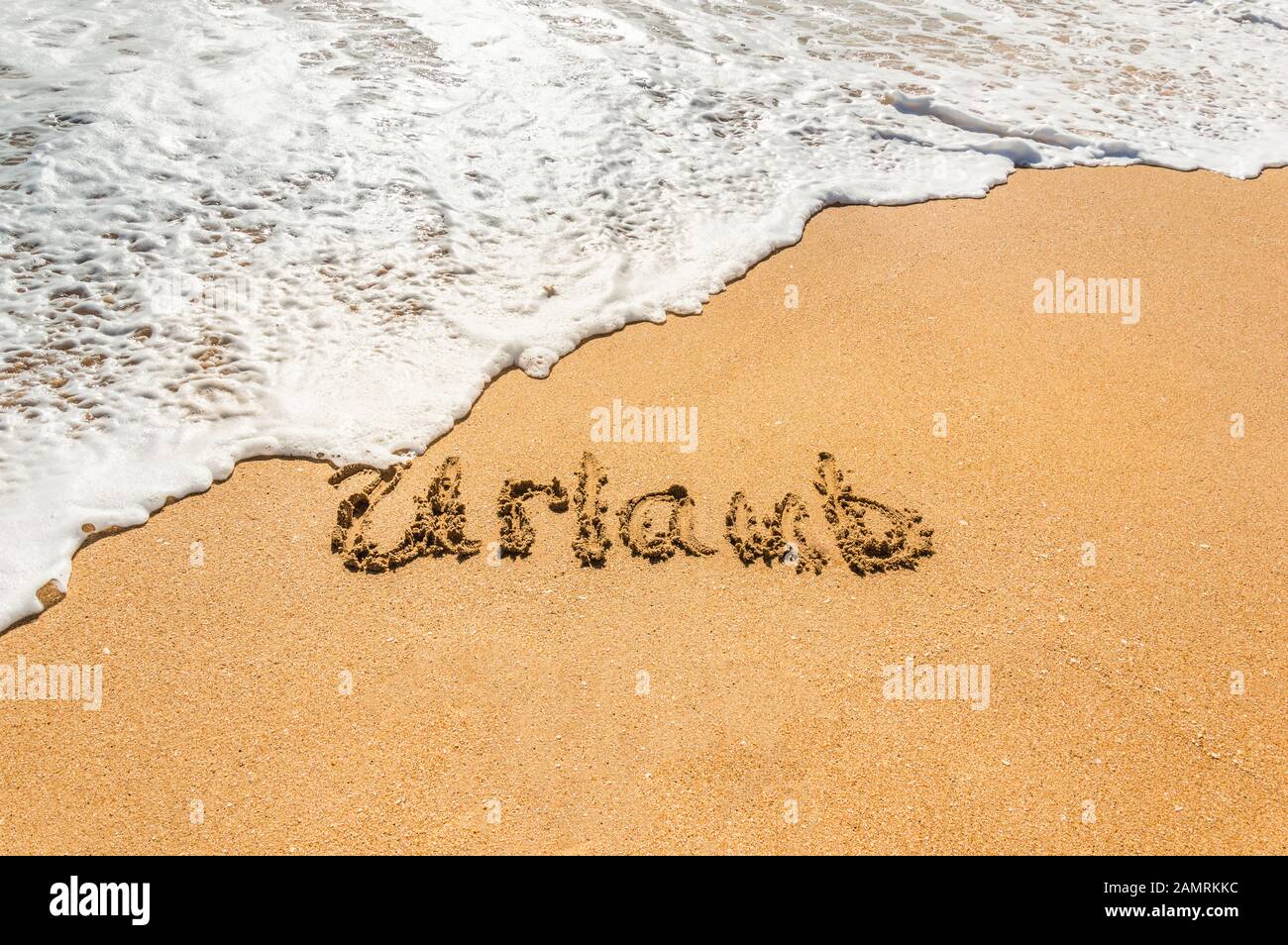Hand written text in German Urlaub (English translation holidays) on the golden beach sand with coming wave. Concept of summer holidays and vacations Stock Photo