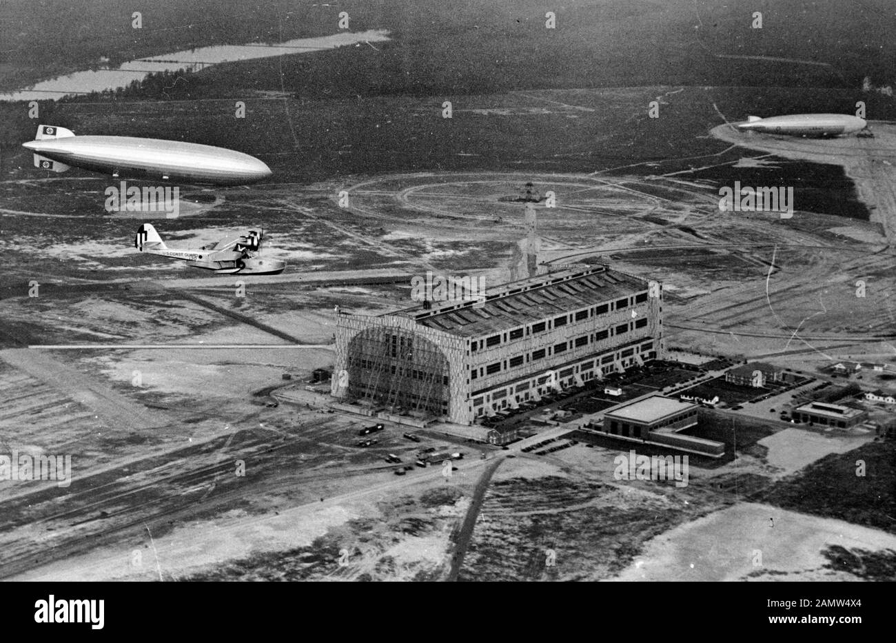 The U.S. Coast Guard Douglas RD-4 Spica (s/n V-125) escorts the German Zeppelin LZ 129 Hindenburg on arrival at Lakehurst, New Jersey (USA), after its inaugural flight from Friedrichshafen, Germany, on the early morning of 9 May 1936. The decommissioned U.S. Navy airship USS Los Angeles (ZR-3) is visible in the background. May 9, 1936 Stock Photo