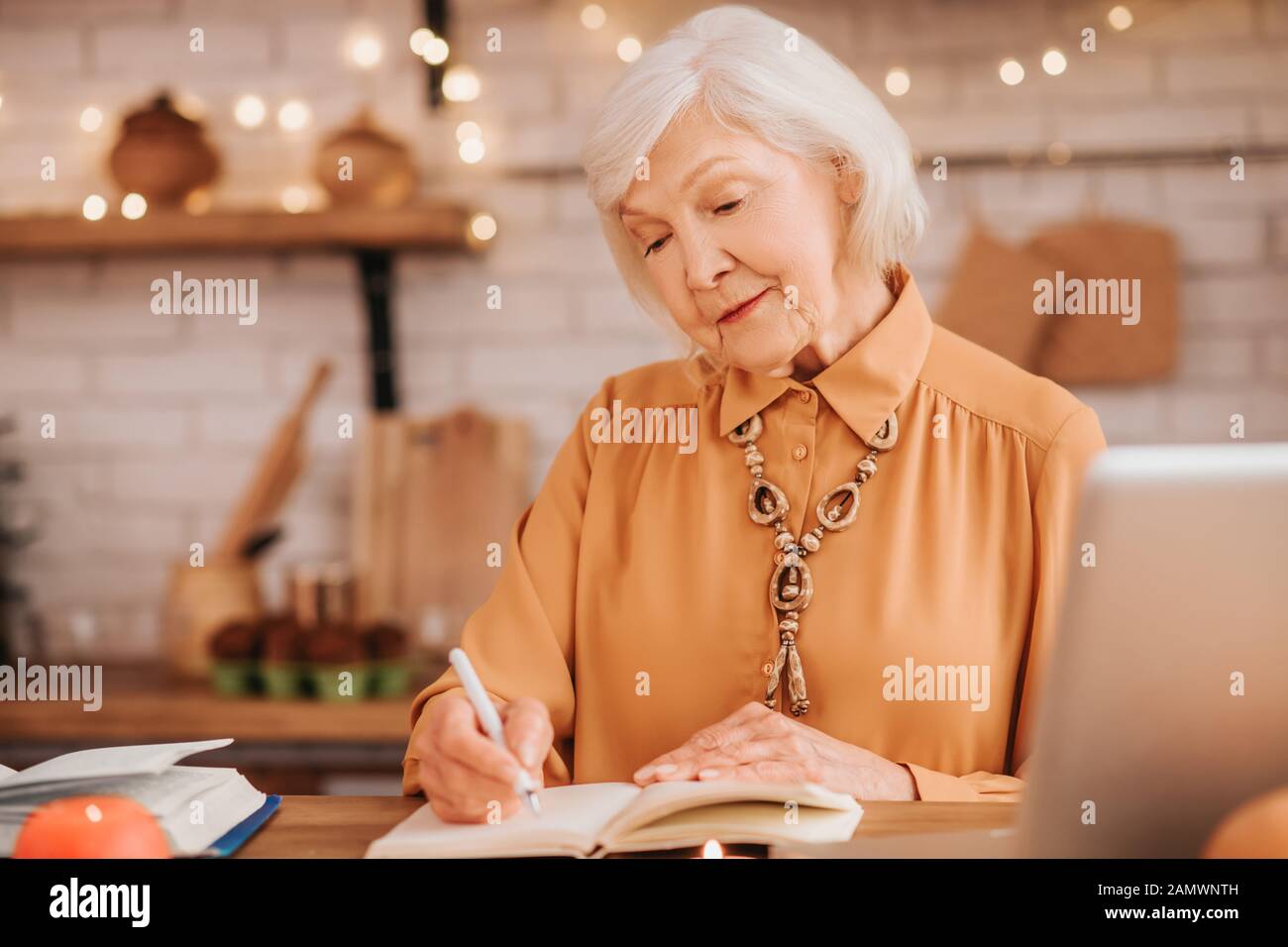 Grey-haired beautiful elderly lady in orange blouse making notes Stock Photo