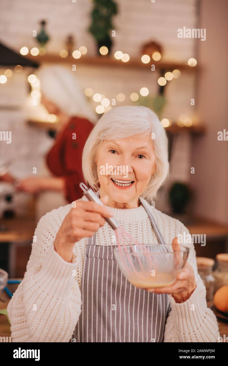 Grey-haired smiling lady in apron whipping milk in a bowl Stock Photo