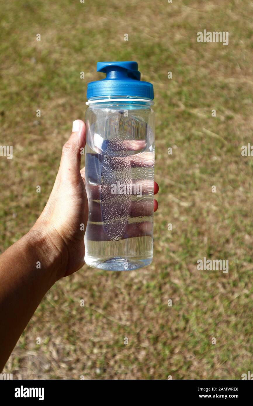 Photo of a hand holding a clear water tumbler at a grassy field on a sunny day. Stock Photo