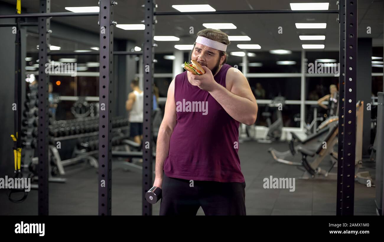 Obese man holding dumbbell eating burger, refusing from doing sport, addiction Stock Photo