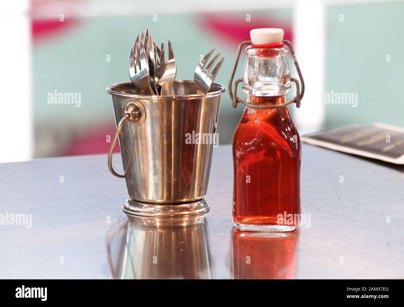 Cutlery in a bronze bucket and a bottle of red grape vinegar on a smooth mirrored table surface at a fish market Stock Photo