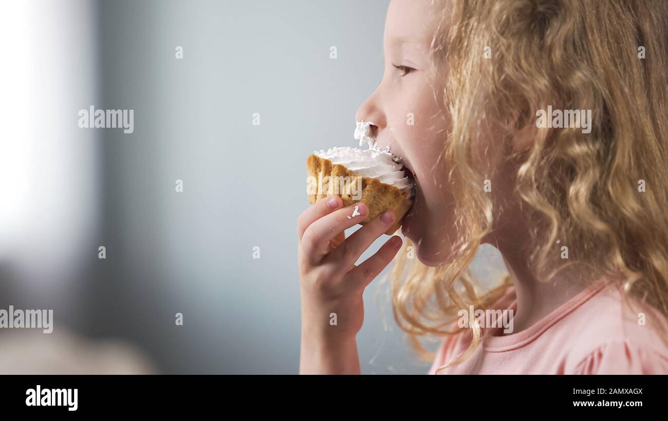 Cute pale preschool girl eating creamy cake and smiling, candy bar at party Stock Photo