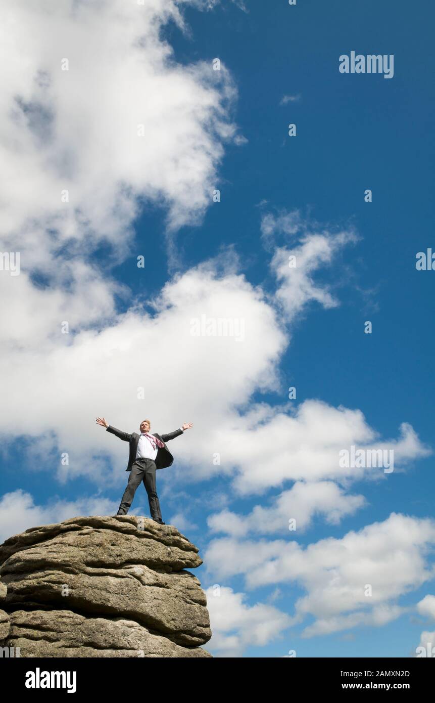 Businessman standing on the top of a distant rocky mountain peak celebrating in blue sky copy space Stock Photo