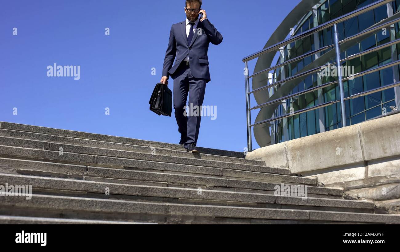 Tired businessman with suitcase talking on phone with partner, going down stairs Stock Photo