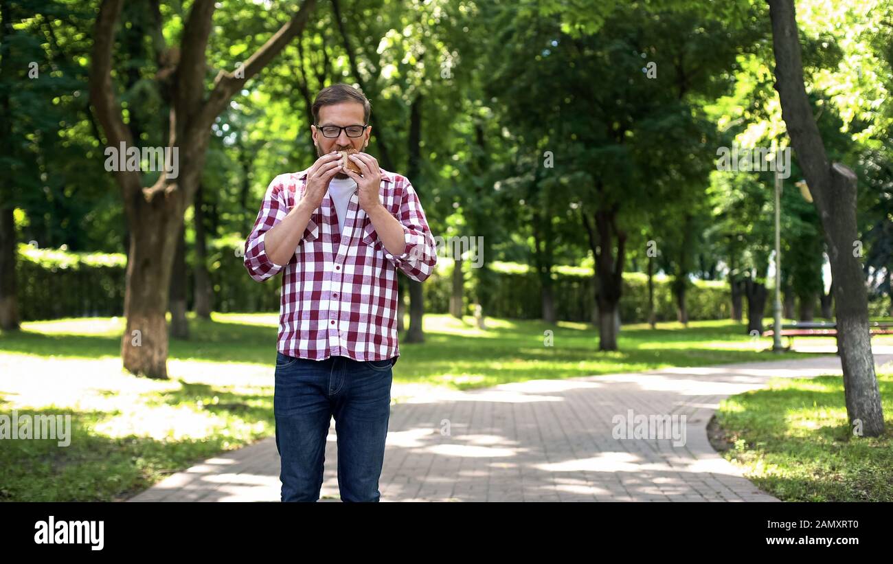 Male eating burger in park, junk food, unhealthy nutrition, busy lifestyle Stock Photo
