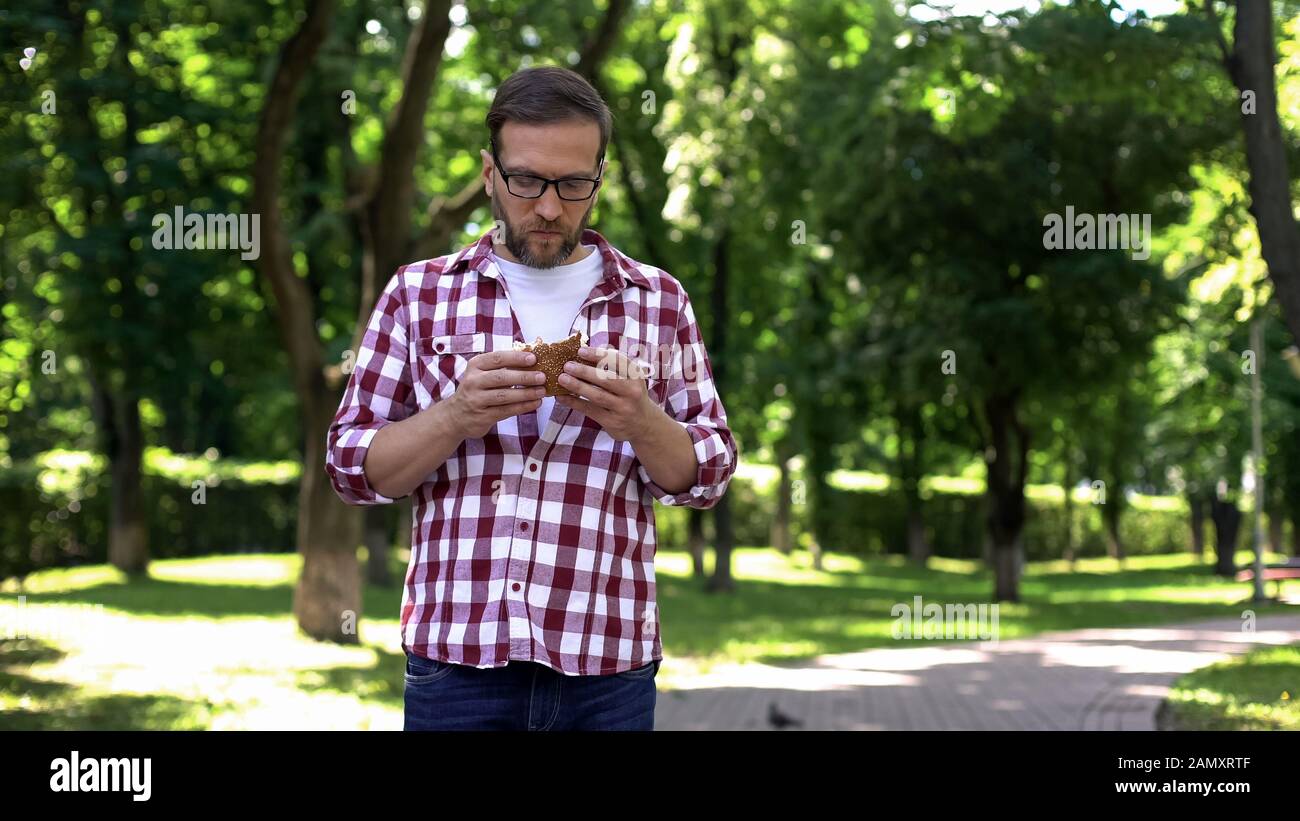 Guy chewing fast food burger in park feeling nausea, food poisoning symptom Stock Photo