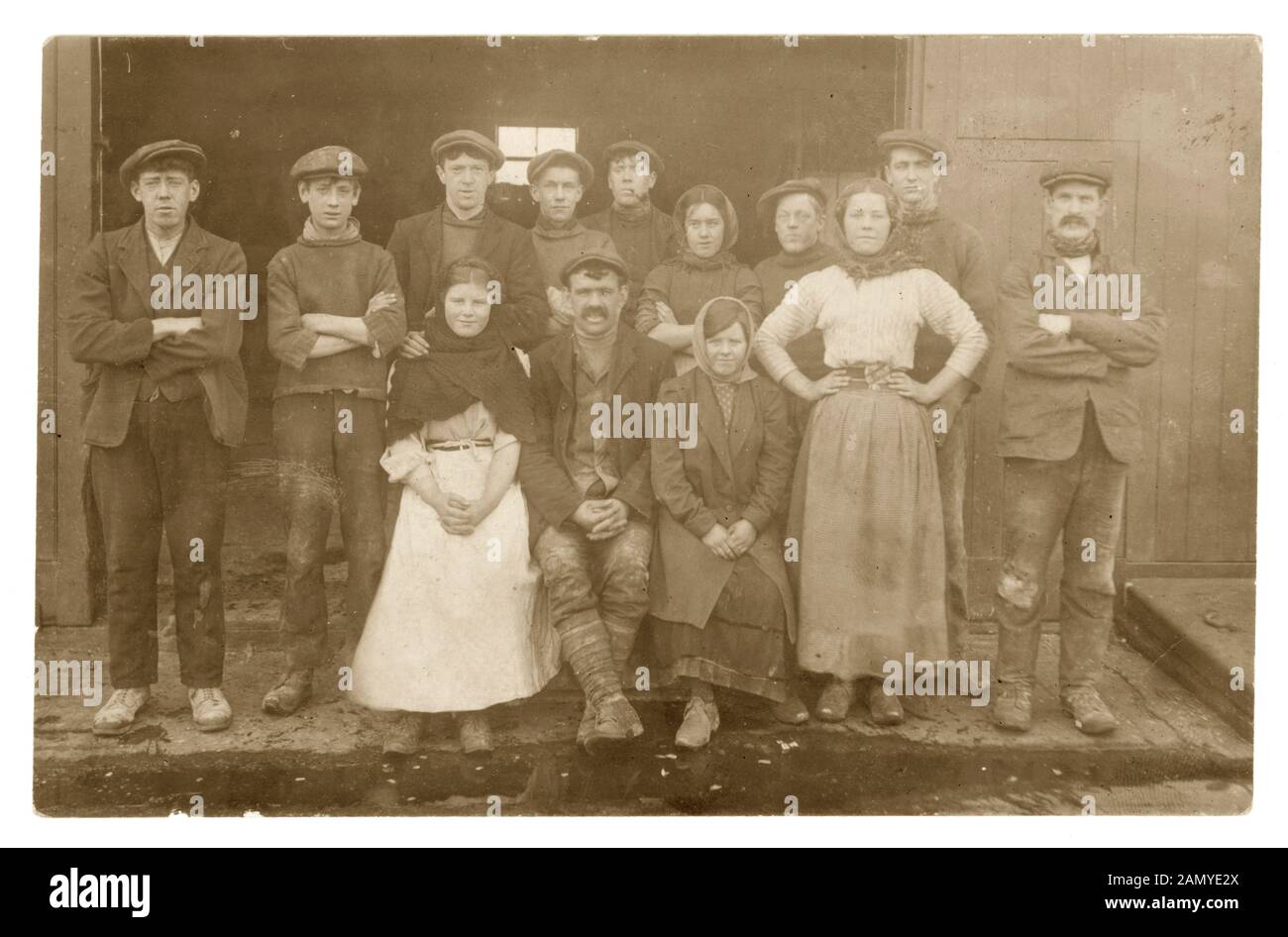 Edwardian photograph of group of young girls and boys with older men, possibly overseers,dressed in work clothes - clogs, shawls. These are probably mill workers as a girl has a belt with scissors in them seen in other mill worker photos possibly Lancashire, England, U.K., circa 1910 Stock Photo