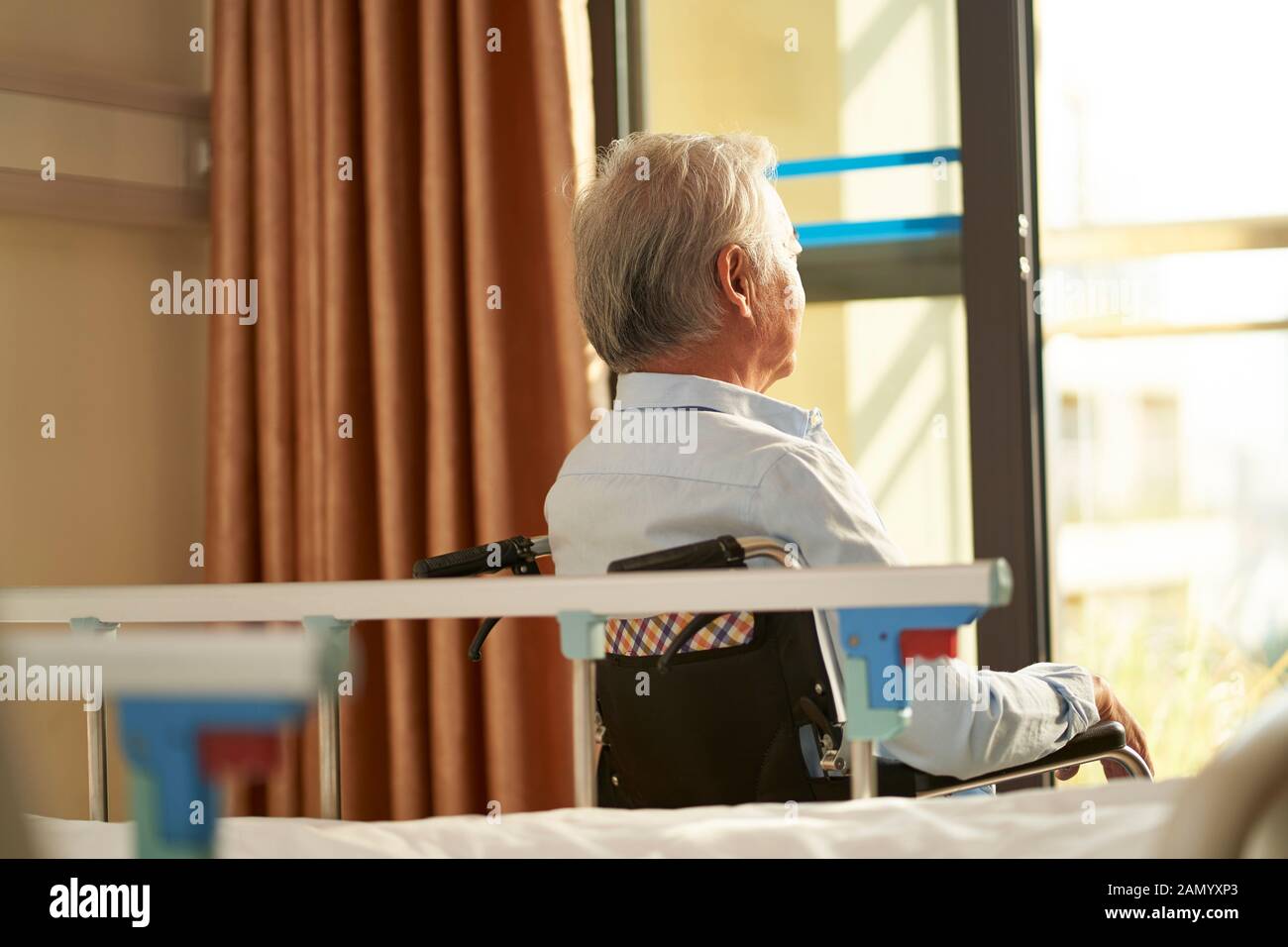 rear view of senior asian man sitting in wheel chair in nusing home or hospital ward looking out of window Stock Photo