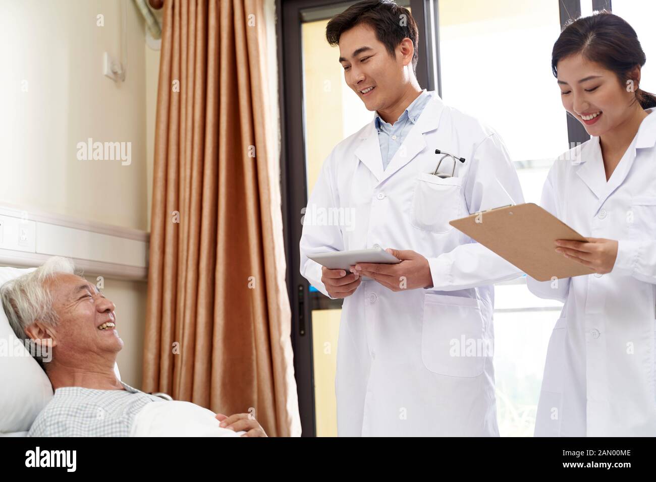 two young friendly asian doctors talking to senior asian male patient at bedside in hospital Stock Photo