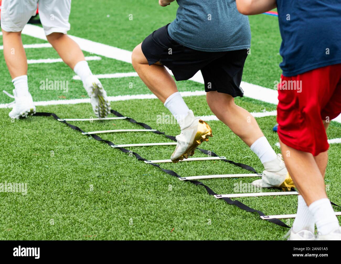 An agility ladder is on a green turf field with three young boys in cleats running through each leg. Stock Photo