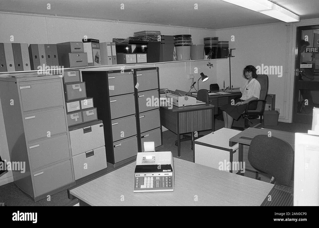 1986, historical, a female clerical worker at a desk in an low-ceiling office, surrounded office supplies and stacks of metal filing cabinets, England, UK. Stock Photo