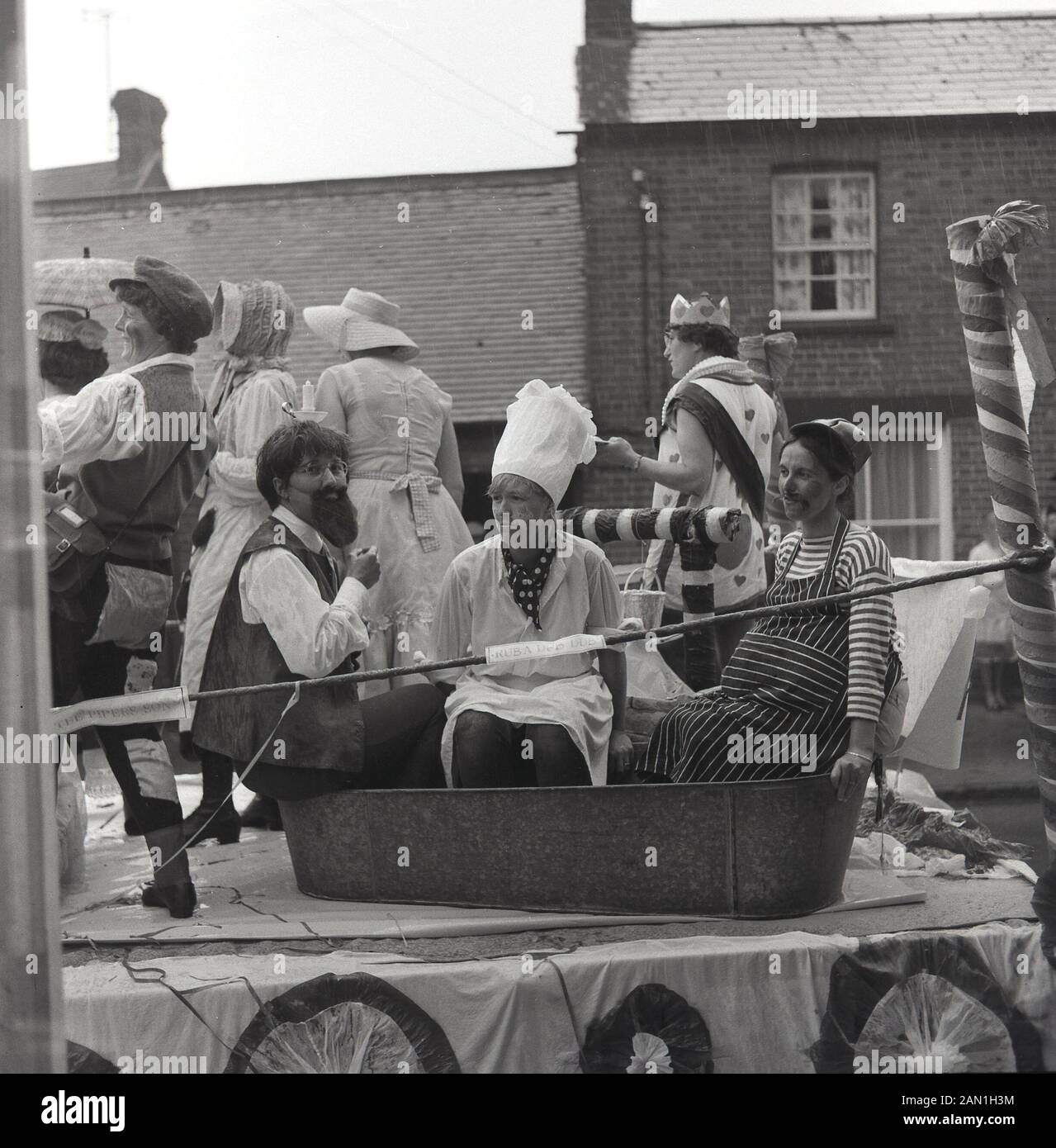 1965, historical, 'Ruba Dub Dub', young adult in costumes sitting in a metal or tin bath on an open-top trailer on a road in a  village carnival, England, UK. Stock Photo