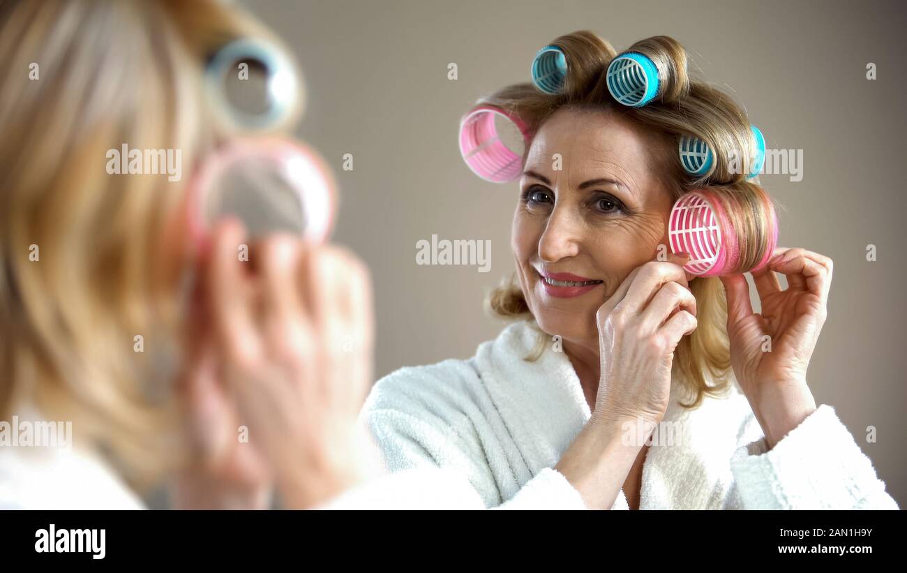 Beautiful old lady with curlers smiling into mirror, enjoying her look, beauty Stock Photo