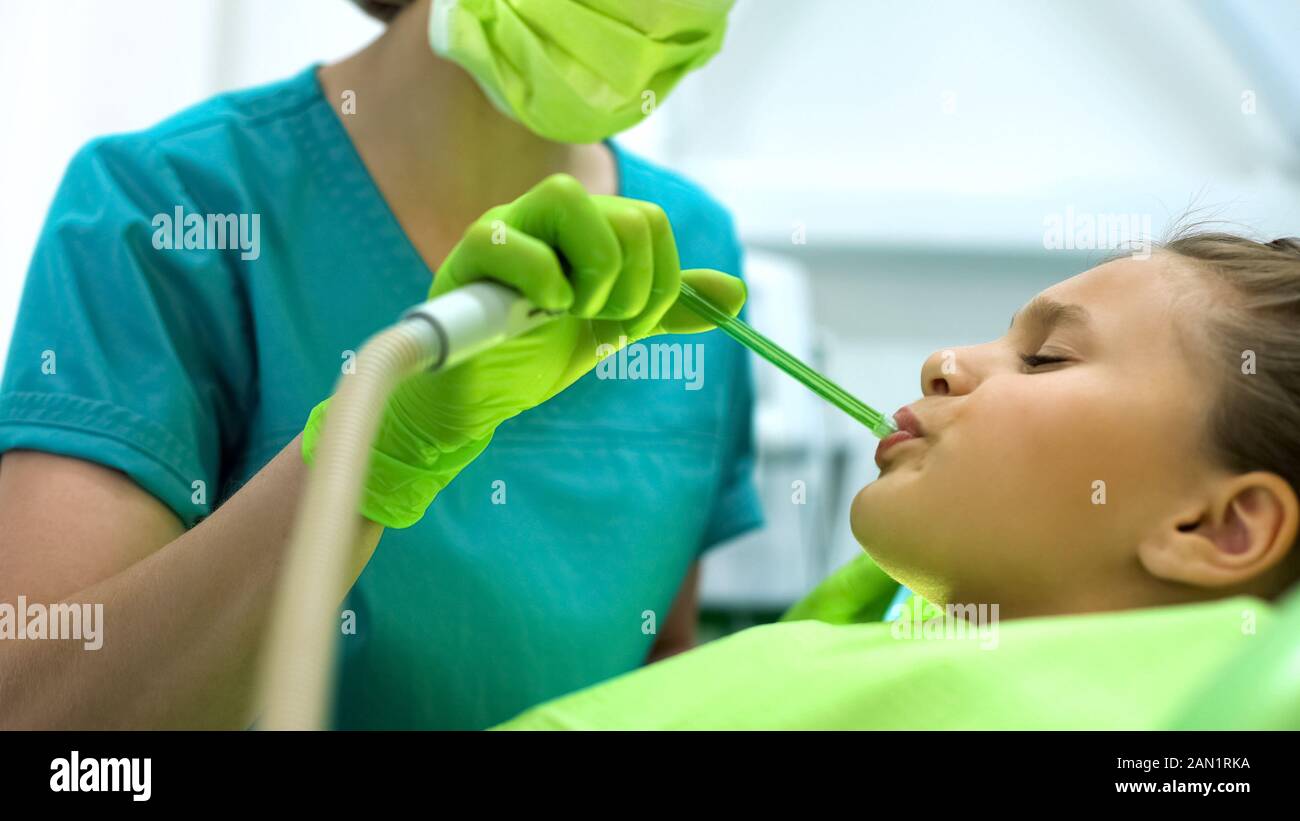 Female dentist using saliva ejector, teenage girl sitting in chair